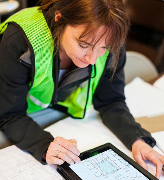 A woman in a green safety vest reviews architectural plans on a tablet using construction management software, with paper blueprints spread out on the table in front of her.
