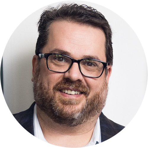 A man with short dark hair, a beard, and glasses smiles at the camera. Wearing a dark jacket over a light shirt, he stands against a plain white background—project-ready like someone using construction management software.