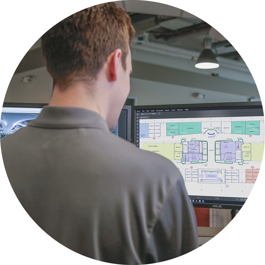 A person with short brown hair, wearing a gray collared shirt, stands at a computer displaying a colorful architectural floor plan on the monitor. The background shows an office interior with exposed ceilings.