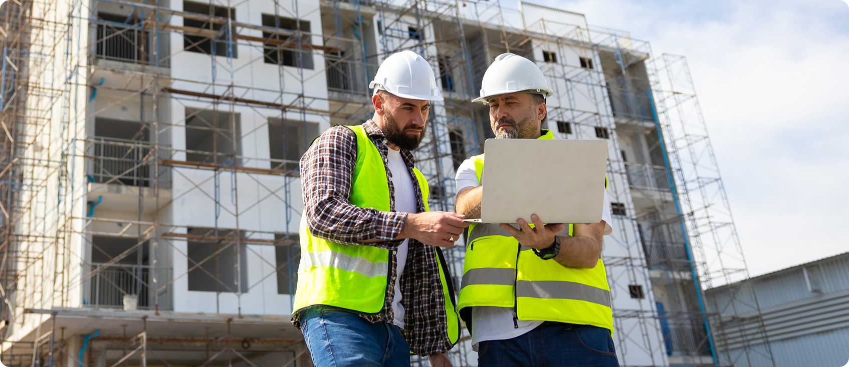  Two construction workers look at laptop together in front of building 